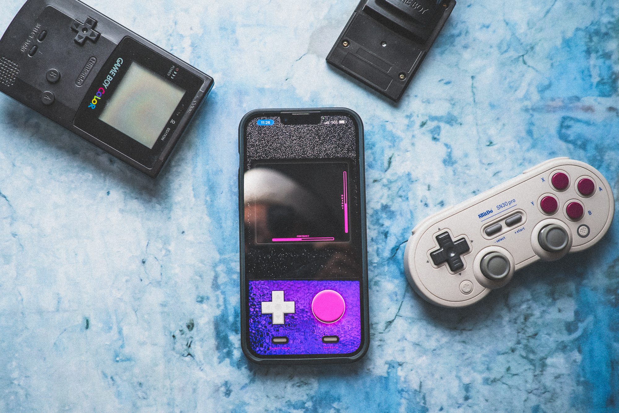 A smartphone with a retro gamepad case, a game controller, and two handheld consoles on a blue surface.
