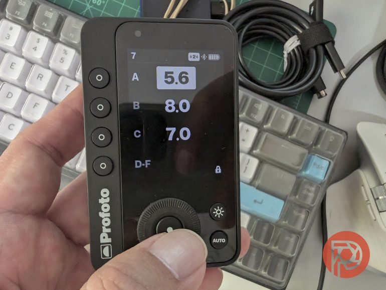 A hand holding a Profoto remote control device with settings displayed; keyboard and cables in background.