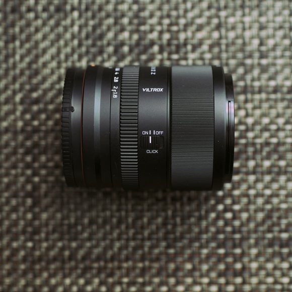A Viltrox camera lens lies on a textured surface with a red circular logo in the bottom right corner.