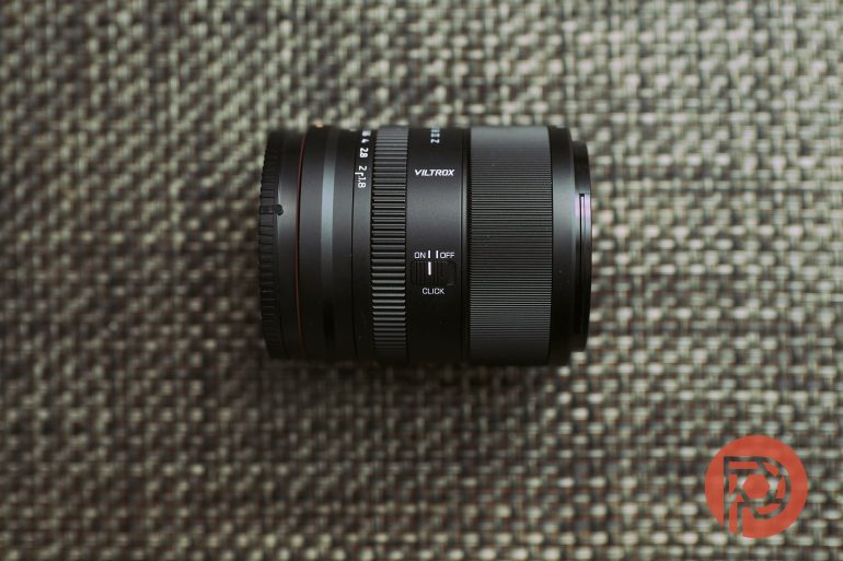 A Viltrox camera lens lies on a textured surface with a red circular logo in the bottom right corner.