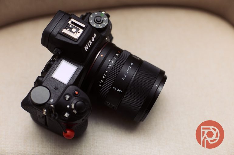 A Nikon camera with a Viltrox 24mm lens rests on a beige surface, with a red logo in the corner.