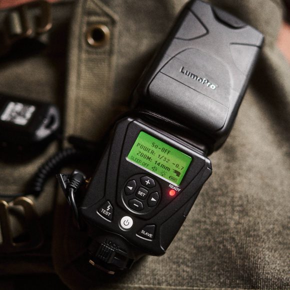 A LumoPro camera flash unit with a digital display, placed on a khaki bag with a buckle.