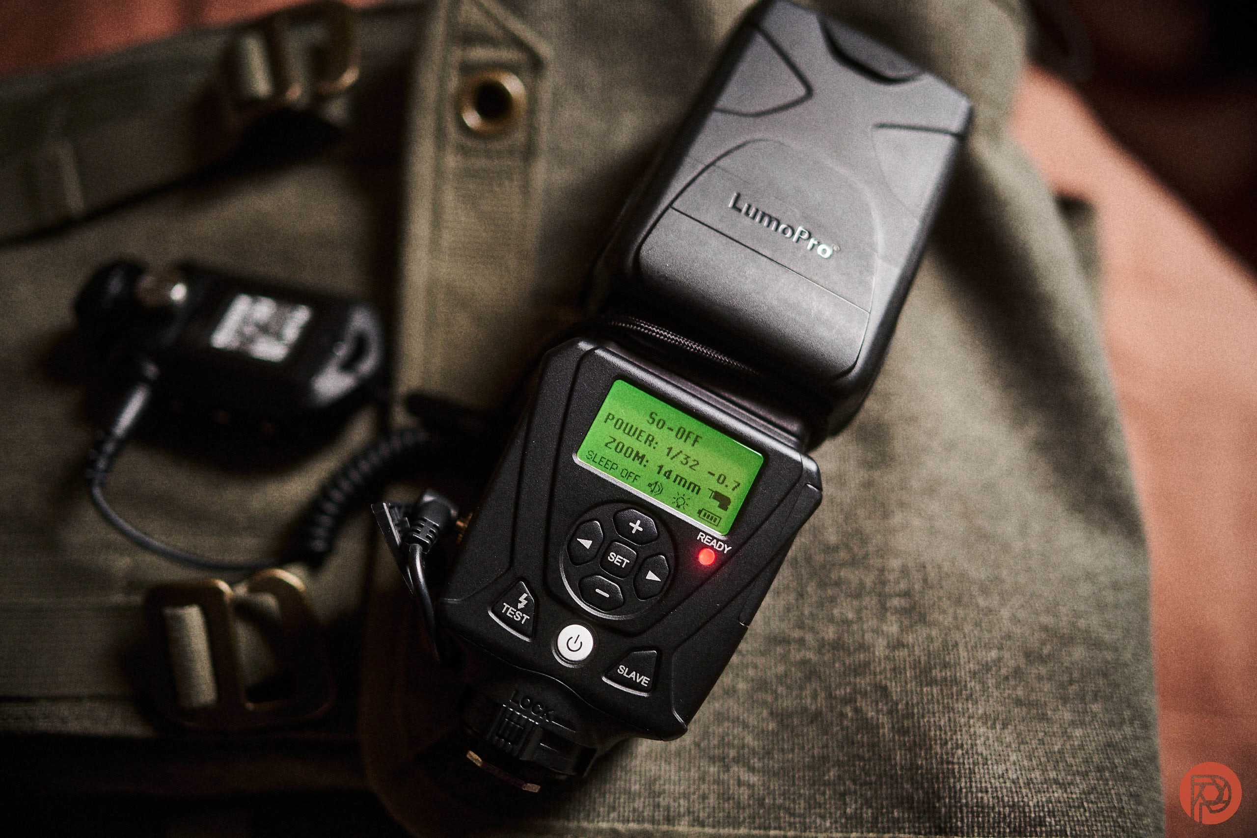 A LumoPro camera flash unit with a digital display, placed on a khaki bag with a buckle.