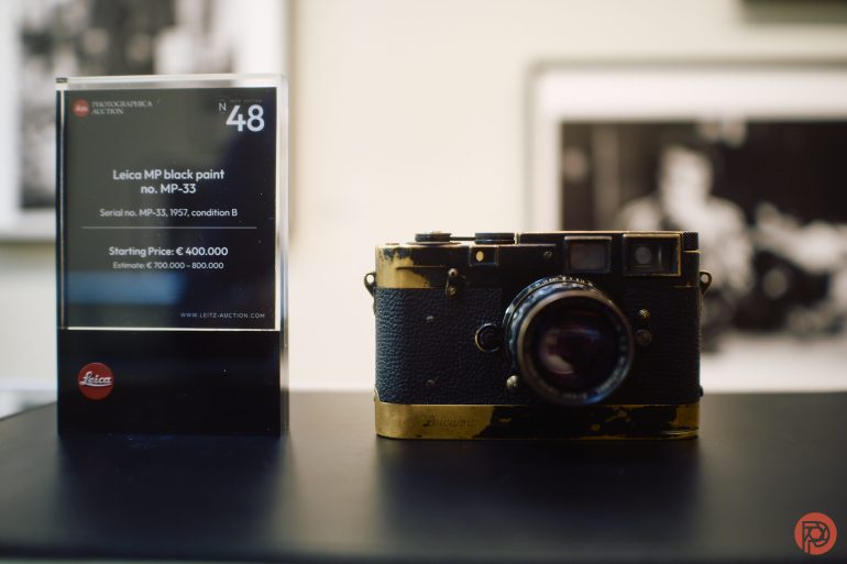 A vintage black Leica MP camera is displayed next to an auction info card on a black surface.