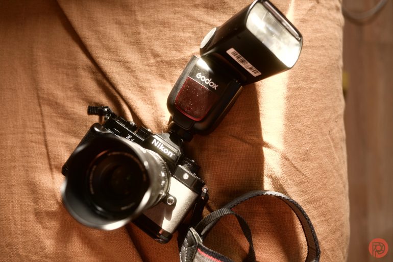 A Nikon camera with a Godox flash attached, resting on a brown fabric surface in bright sunlight.