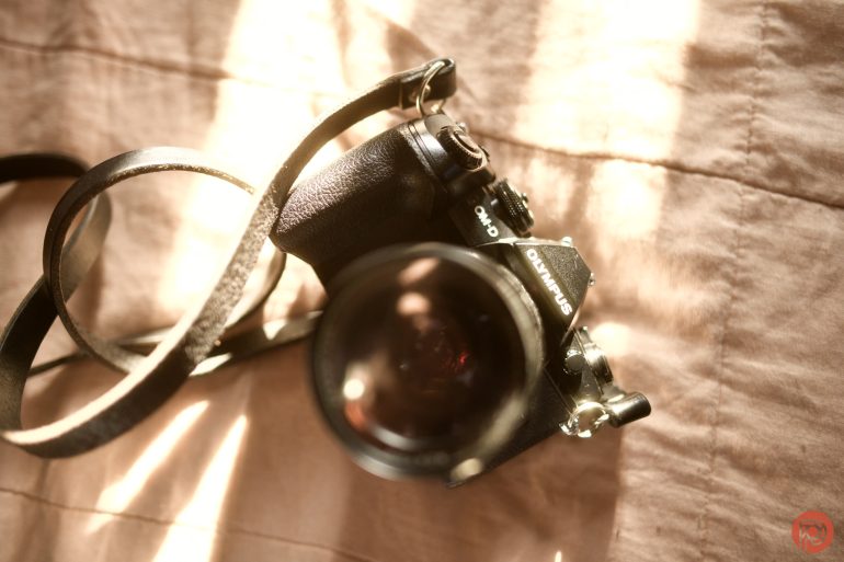 A black Olympus OM-10 film camera with a strap lies on a sunlit, peach-colored fabric surface.