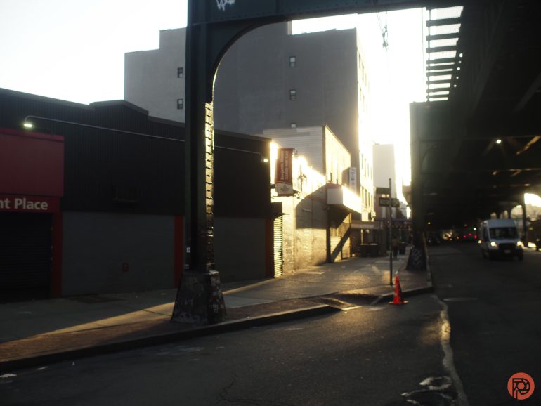 A city street at sunset with a shadowy building, sidewalk, and an elevated train track overhead.
