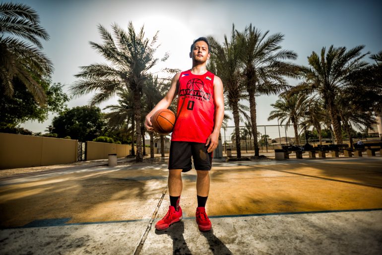 A basketball player in a red jersey stands on an outdoor court holding a basketball, with palm trees behind him.