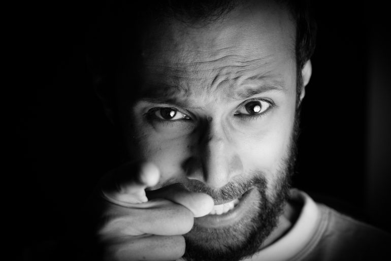 A man with a beard points at the camera, eyebrows furrowed and expression intense, in black and white lighting.