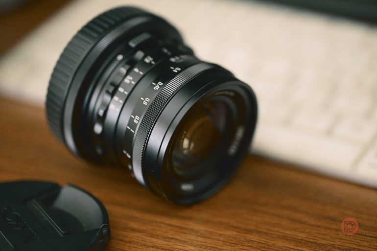 A camera lens resting on a wooden surface near a keyboard and a detached lens cap.