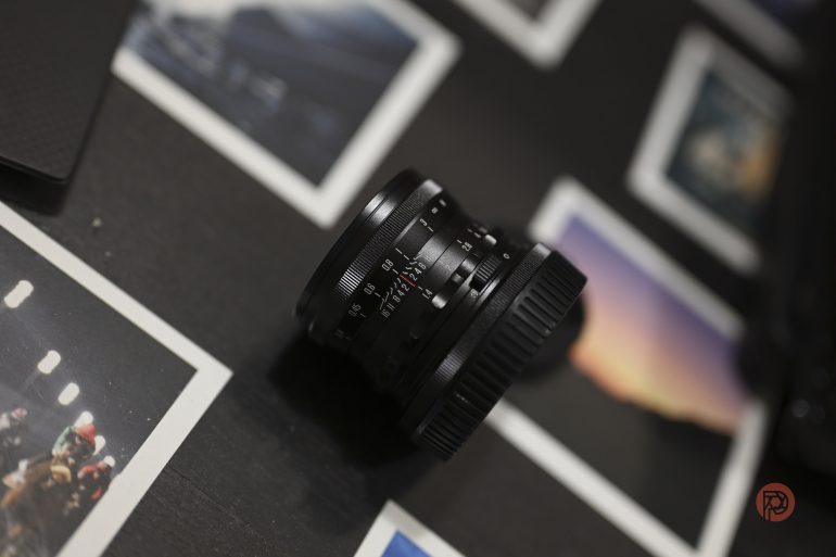 A camera lens rests on a table surrounded by several scattered photographs.