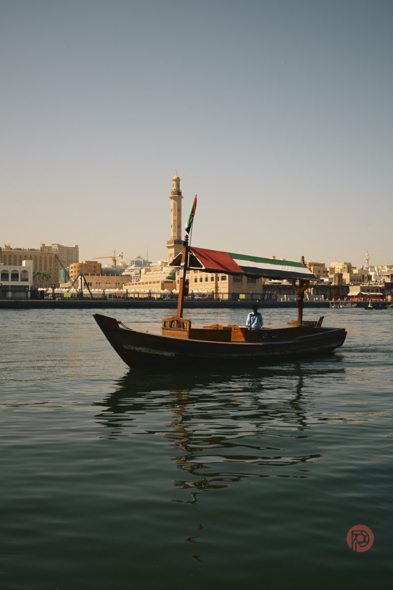A traditional wooden boat with a canopy floats on calm water, city buildings and a mosque in the background.