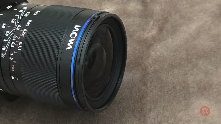 A Laowa camera lens resting on a brown textured surface.