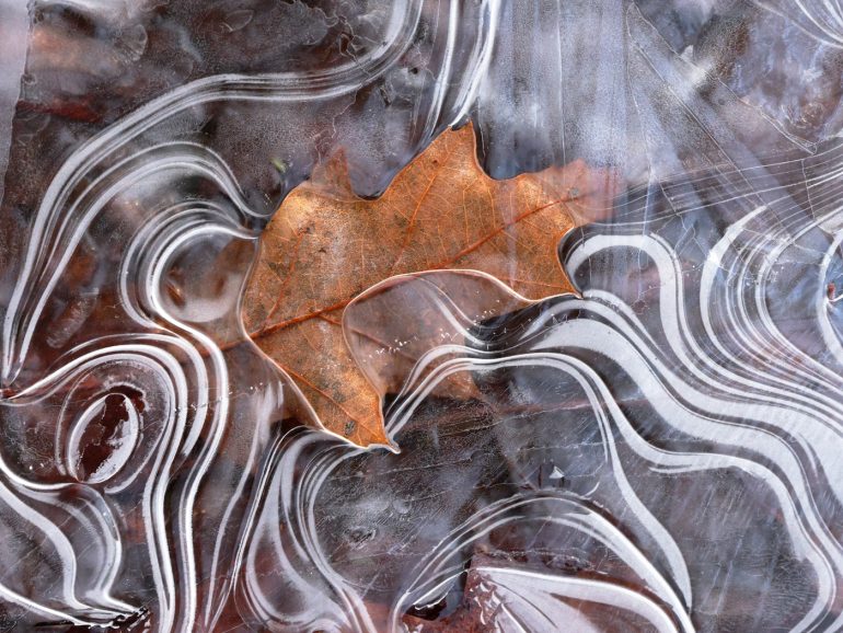 A brown leaf rests on swirling, patterned ice with white lines and frosty textures.