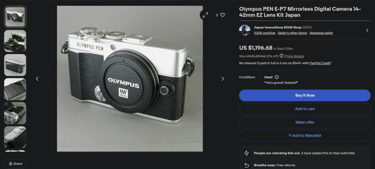 Olympus PEN E-P7 digital camera with attached lens cap on a grey surface, shown from a slight angle.
