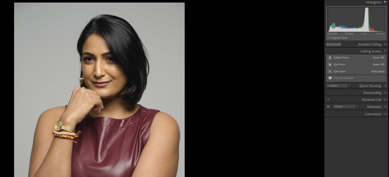 Woman with short black hair in a sleeveless maroon top, smiling with hand on chin, grey background.