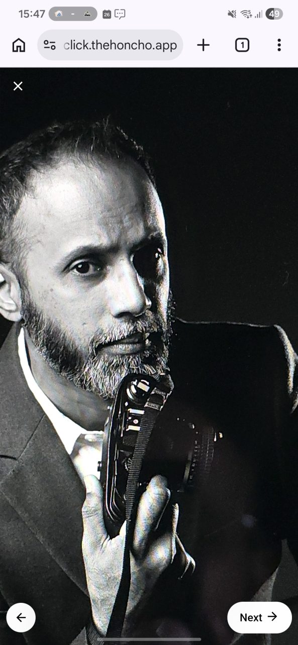 Black and white photo of a man with a beard holding a camera, looking seriously at the viewer.