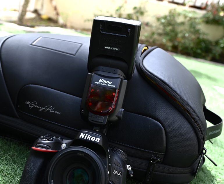 A Nikon DSLR camera with a Speedlight flash sits on a black camera bag on green grass.