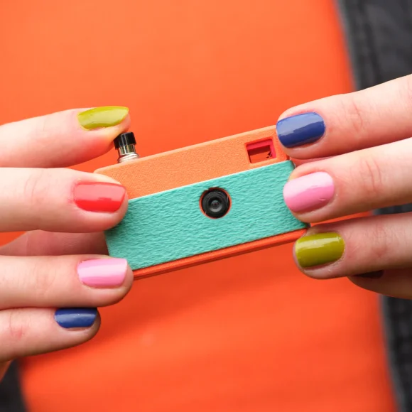 Close-up of hands with colorful nails holding a small, retro-style orange and teal camera.