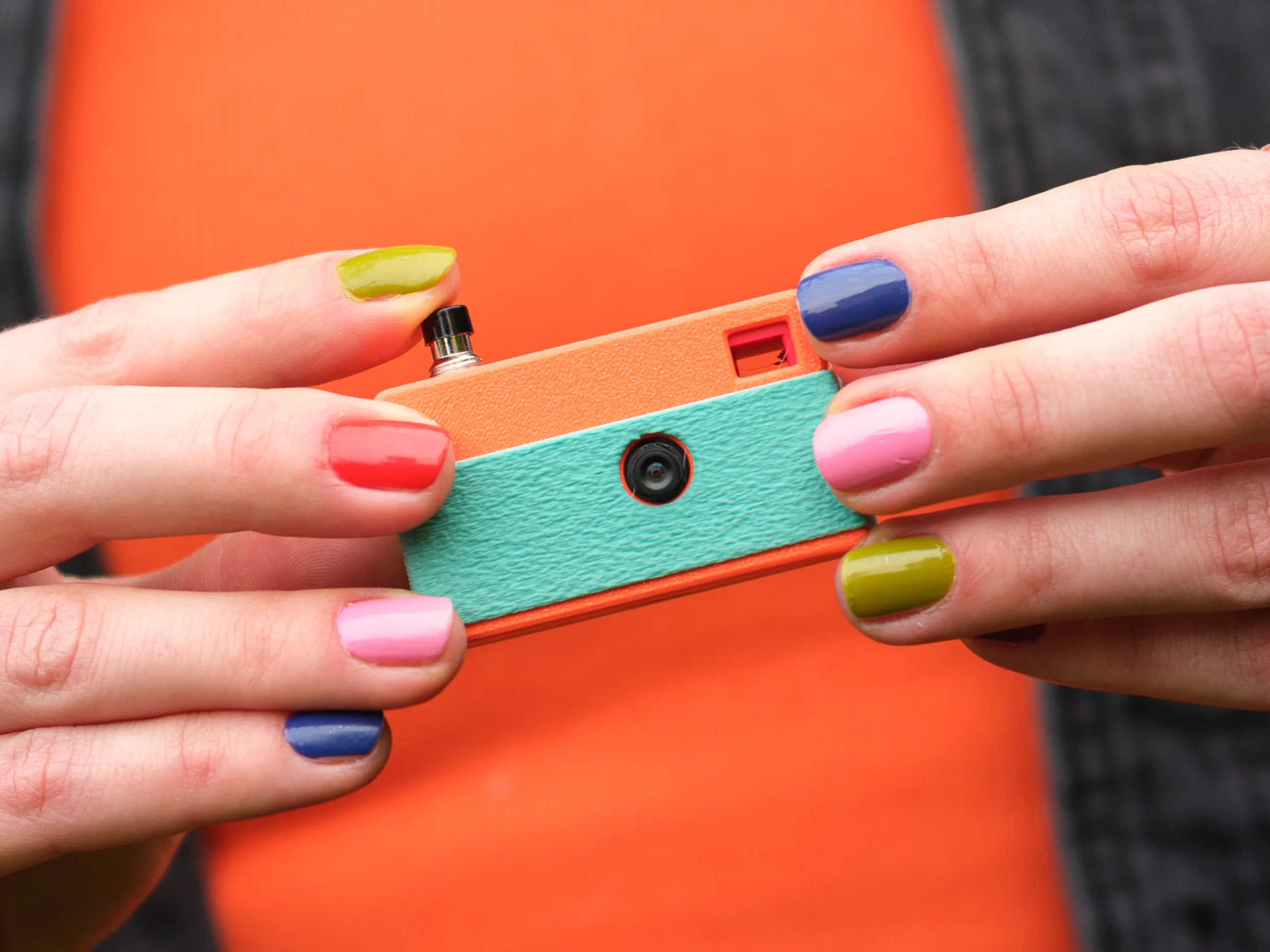 The Keymera is the Cutest Key to a Photographer's Heart Close-up of hands with colorful nails holding a small, retro-style orange and teal camera.