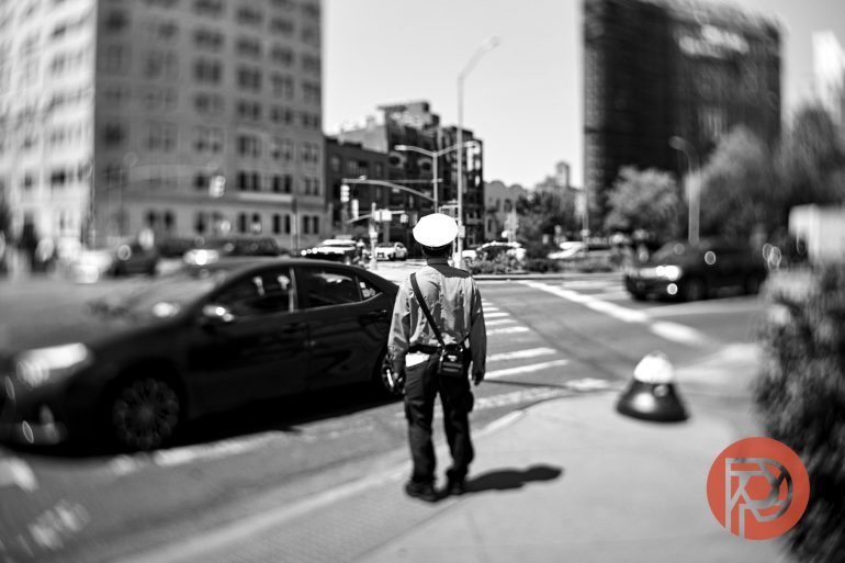 A traffic officer stands at a crosswalk in a city intersection, with blurred buildings and cars in the background.