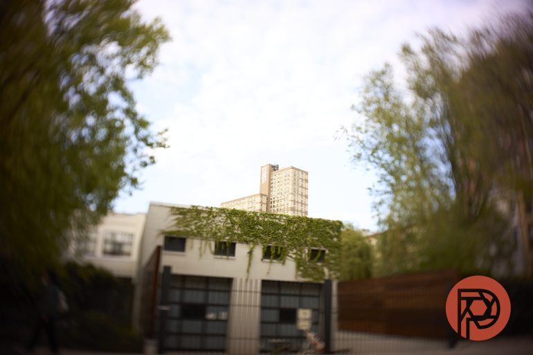 A blurred view of a white building with ivy, tall apartments behind, and trees on both sides; red logo at bottom right.