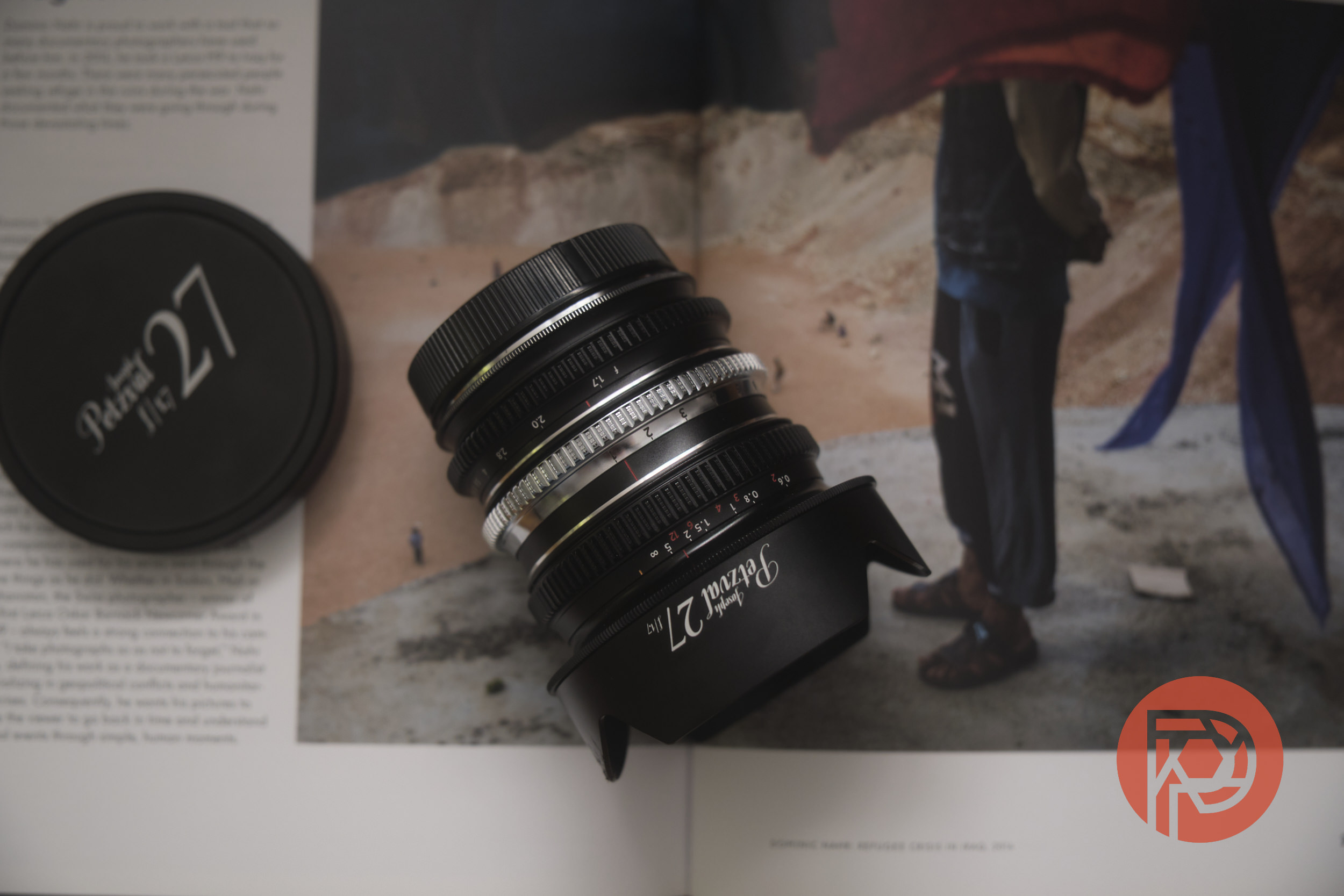 Lomography Joseph Petzval 27 f1.7 Review: Swirly Bokeh is Nice, but Something's Missing Close-up of a camera lens and lens cap resting on an open magazine with a photo of a person outdoors.