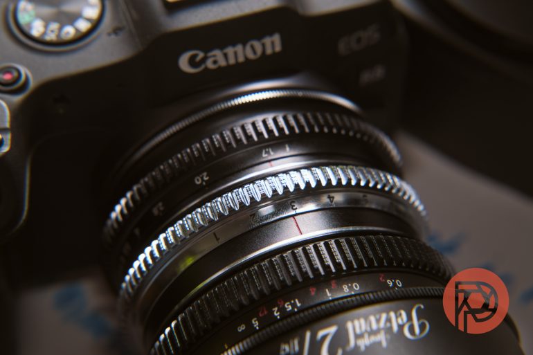 Close-up of a Canon camera lens with visible focus and aperture markings, and a red logo in the corner.