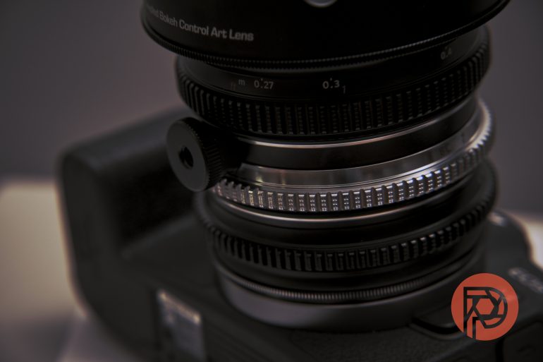 Close-up of a camera lens with adjustment rings and a red logo in the bottom right corner.