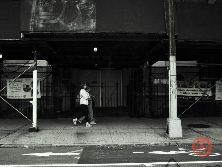 Two people walk past a dark, covered sidewalk beneath scaffolding in an urban setting.