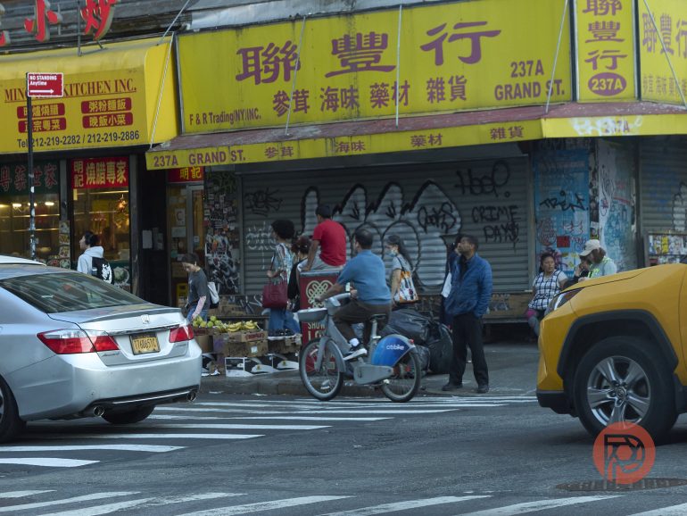 People browsing goods on a busy Chinatown sidewalk with yellow storefronts and cars passing by.