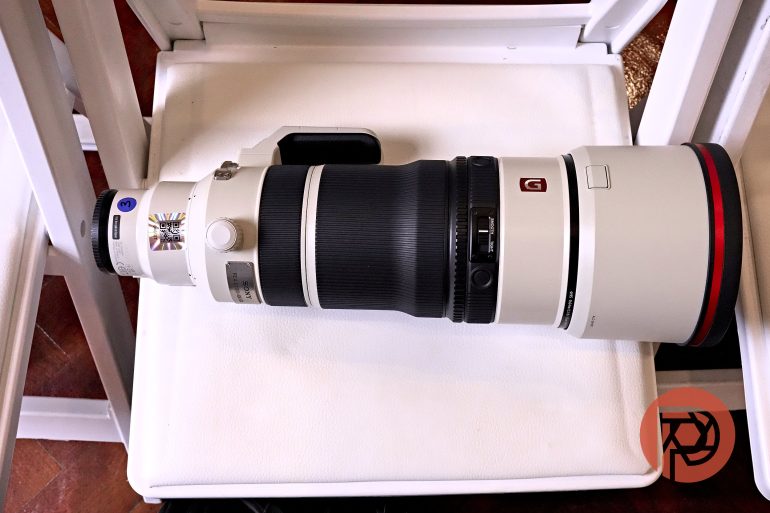 A large Sony telephoto camera lens lies on a white surface, with a red logo in the bottom right corner.