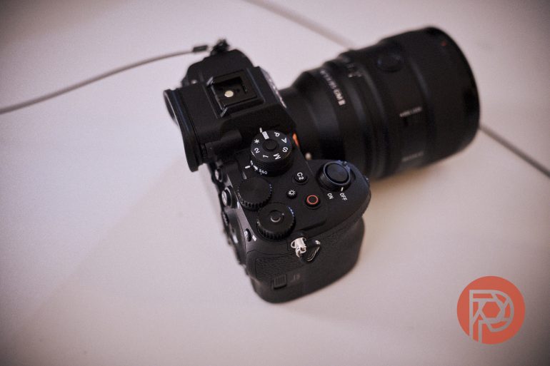 Black digital camera with lens resting on a white surface, red logo in the bottom right corner.