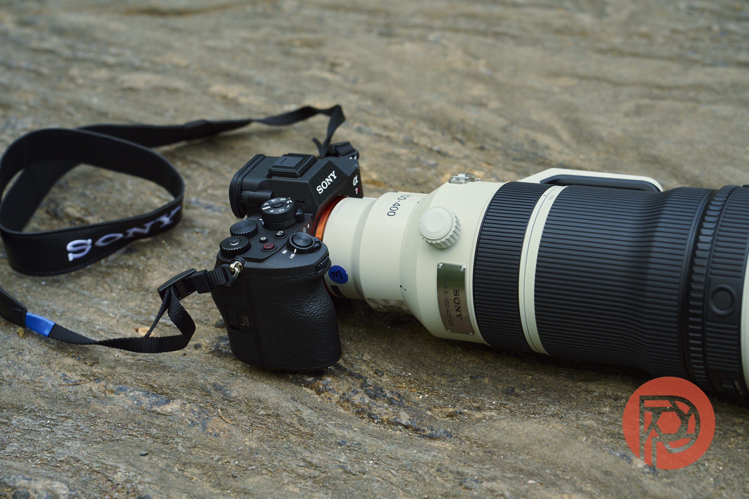 A Sony camera with a large telephoto lens rests on a textured stone surface; red logo at bottom right.