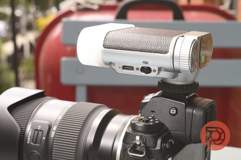 A close-up of a digital camera with a mounted microphone, outdoors, in front of a red background.