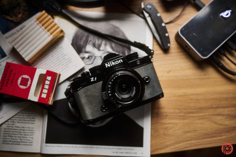 Nikon camera, cigarettes, and a folder on a wooden table.
