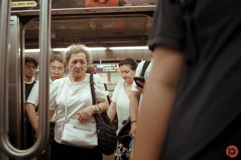 Crowded NYC subway platform with passengers waiting to board, including an older woman in a white top holding a purse.