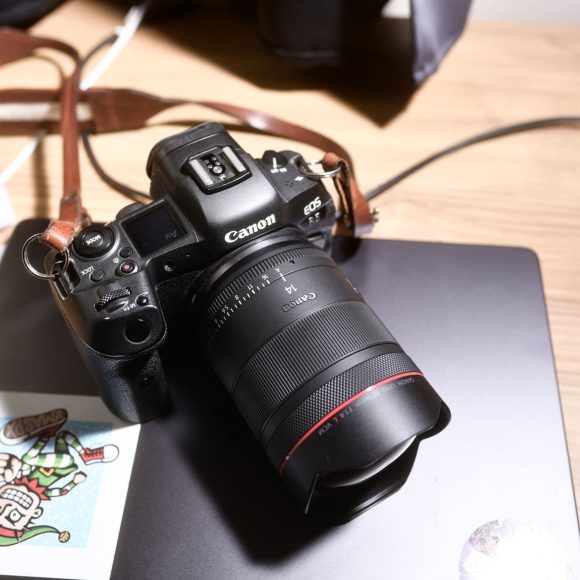 A Canon camera with a lens sits on a closed laptop, next to a sticker and a smartphone on a desk.