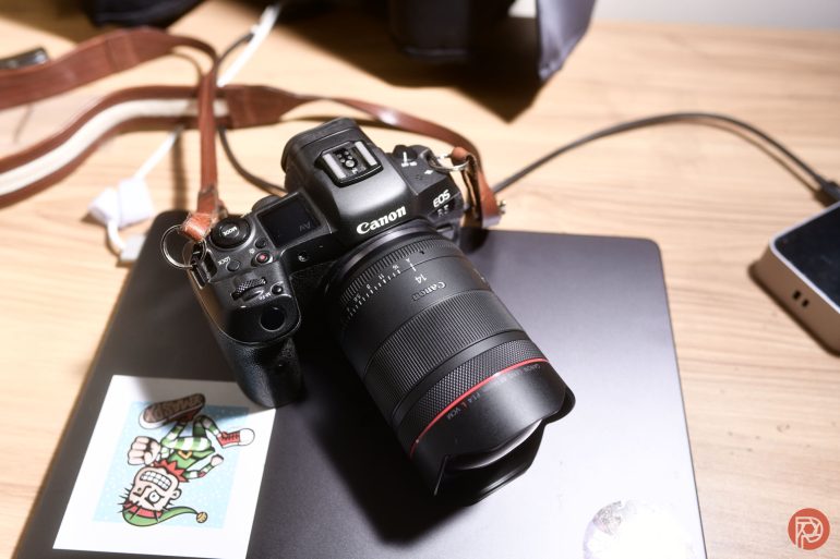A Canon camera with a lens sits on a closed laptop, next to a sticker and a smartphone on a desk.