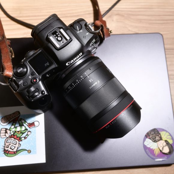 A Canon camera rests on a closed laptop with two colorful stickers on a wooden desk.