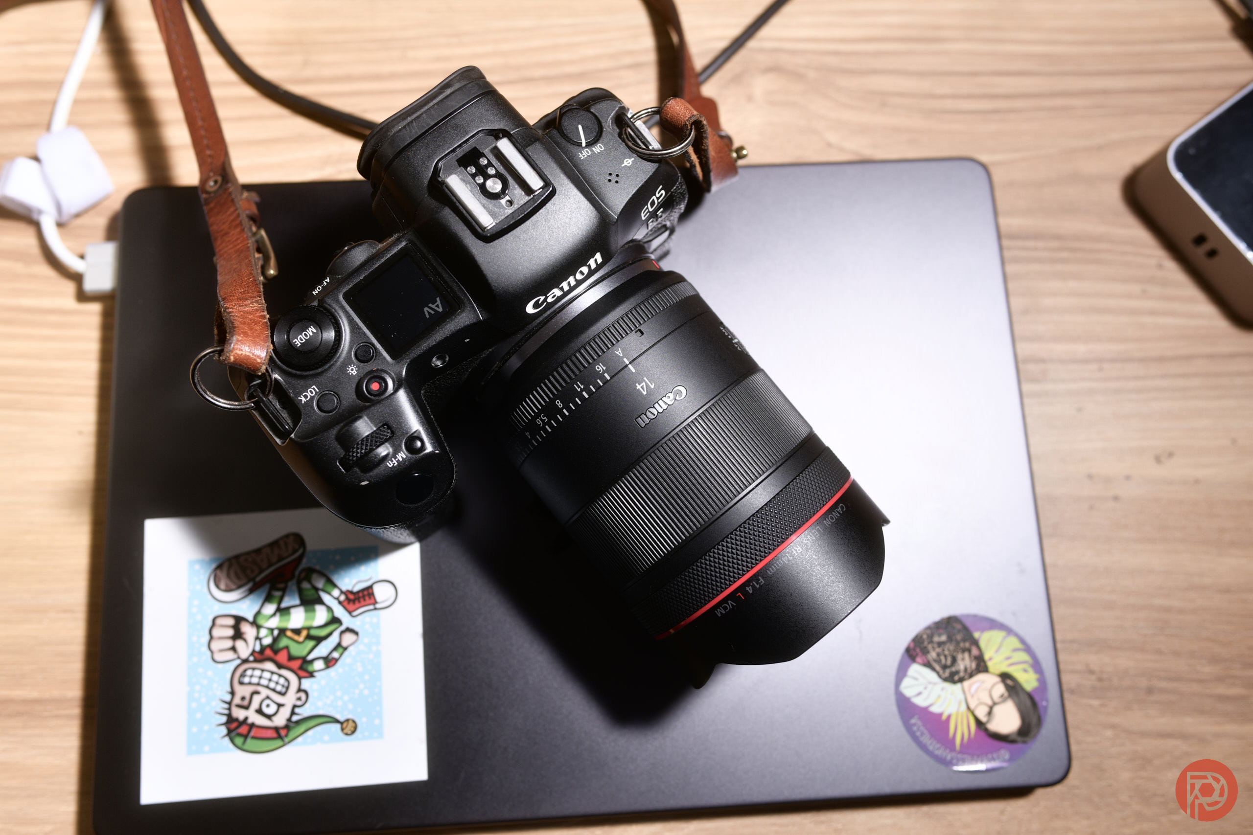 A Canon camera rests on a closed laptop with two colorful stickers on a wooden desk.