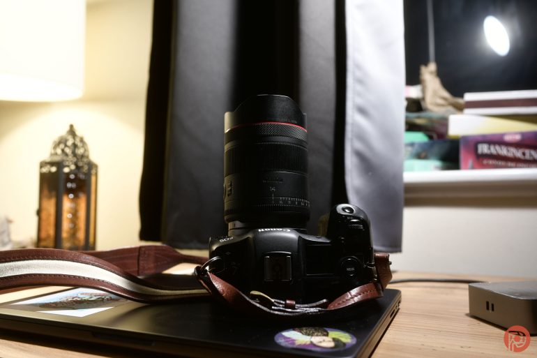 A Canon camera with a leather strap rests on a closed laptop near a window and some stacked books.