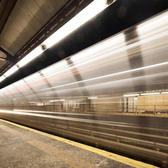 A subway train speeds through a dimly lit station with a 52 St sign and empty platform.