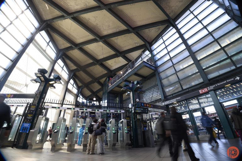 People enter and exit through turnstiles at a busy, modern subway station with high ceilings and glass walls.