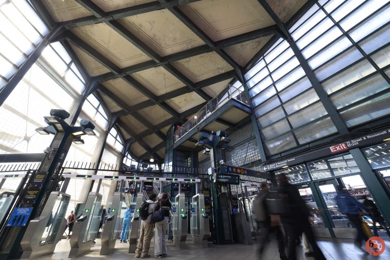 Commuters pass through turnstiles in a spacious, sunlit subway station with high ceilings and large windows.