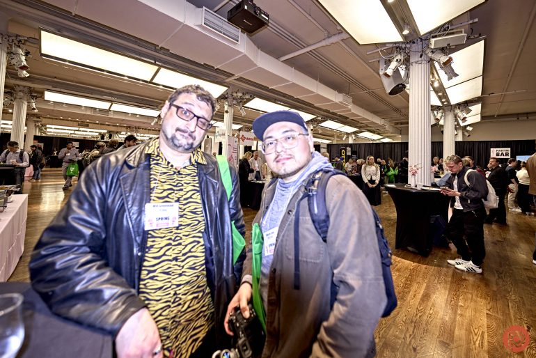 Two men posing together at an indoor event with people and tables in the background.