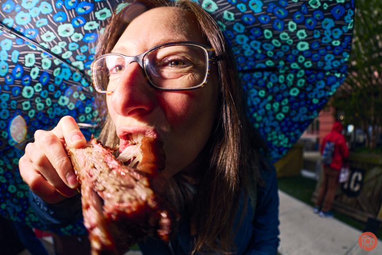 Close-up of a woman in glasses biting into a large piece of barbecued meat under a floral umbrella.