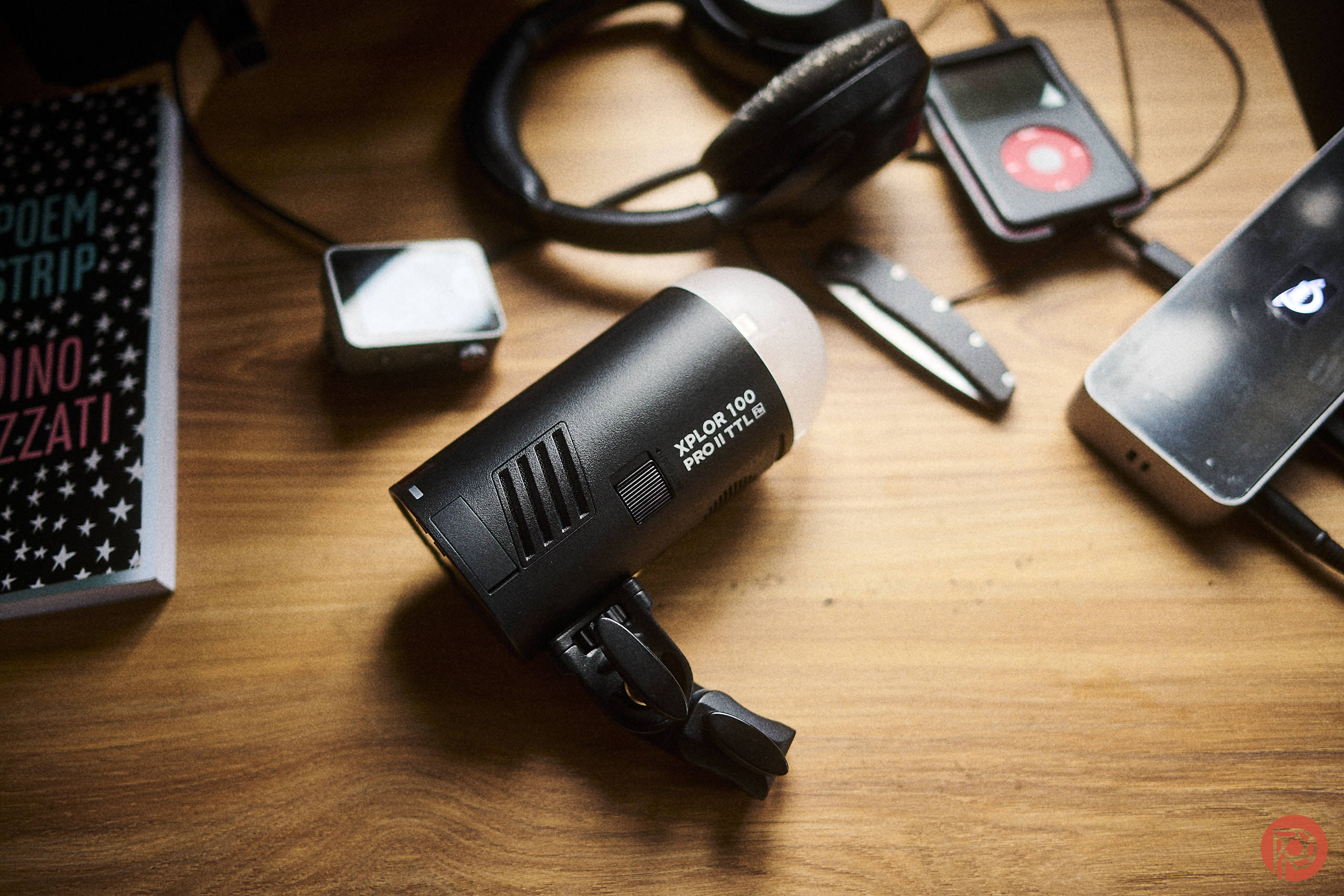The Flashpoint XPLOR100 Pro II TTL is a Joy to Use A portable strobe light sits on a wooden table next to headphones and a media player.