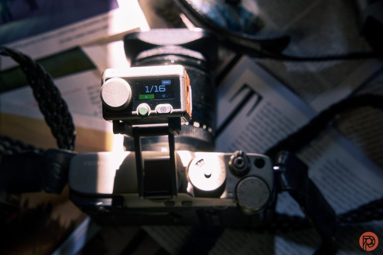 A close-up of a camera with an external flash unit displaying 1/16 on its screen, surrounded by papers.
