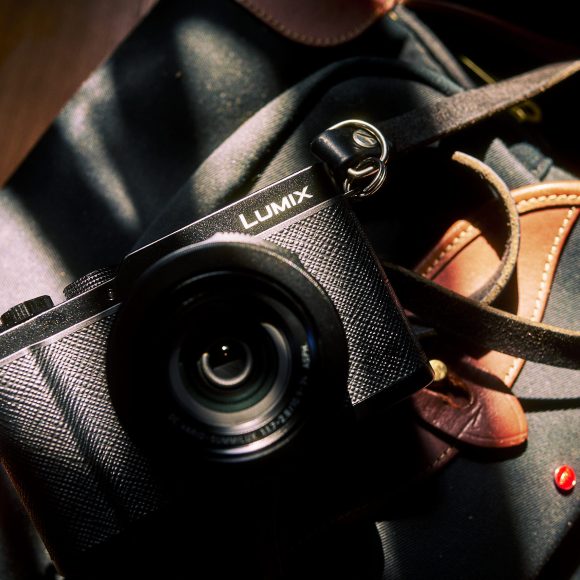 A Lumix camera rests on a dark bag with brown leather accents, lit by a beam of sunlight.